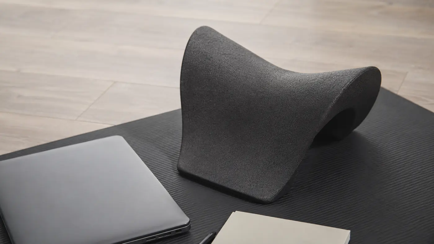 A foam neck stretcher pillow resting on a floor mat, unused, next to a laptop and notebook